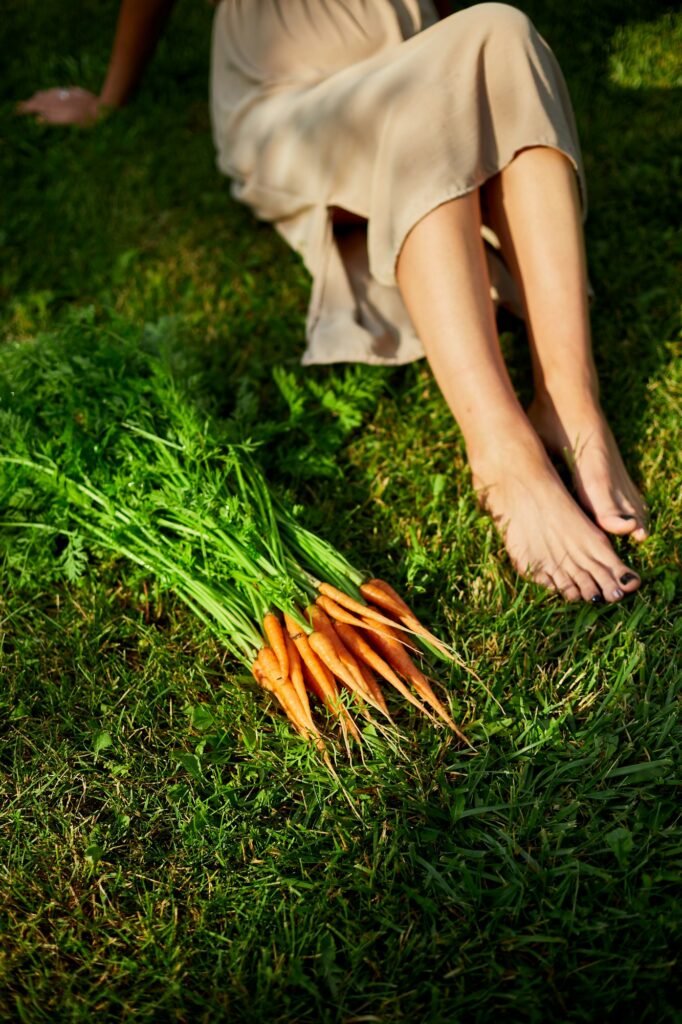 Woman sitting barefoot on grass with branch of raw organic carrots